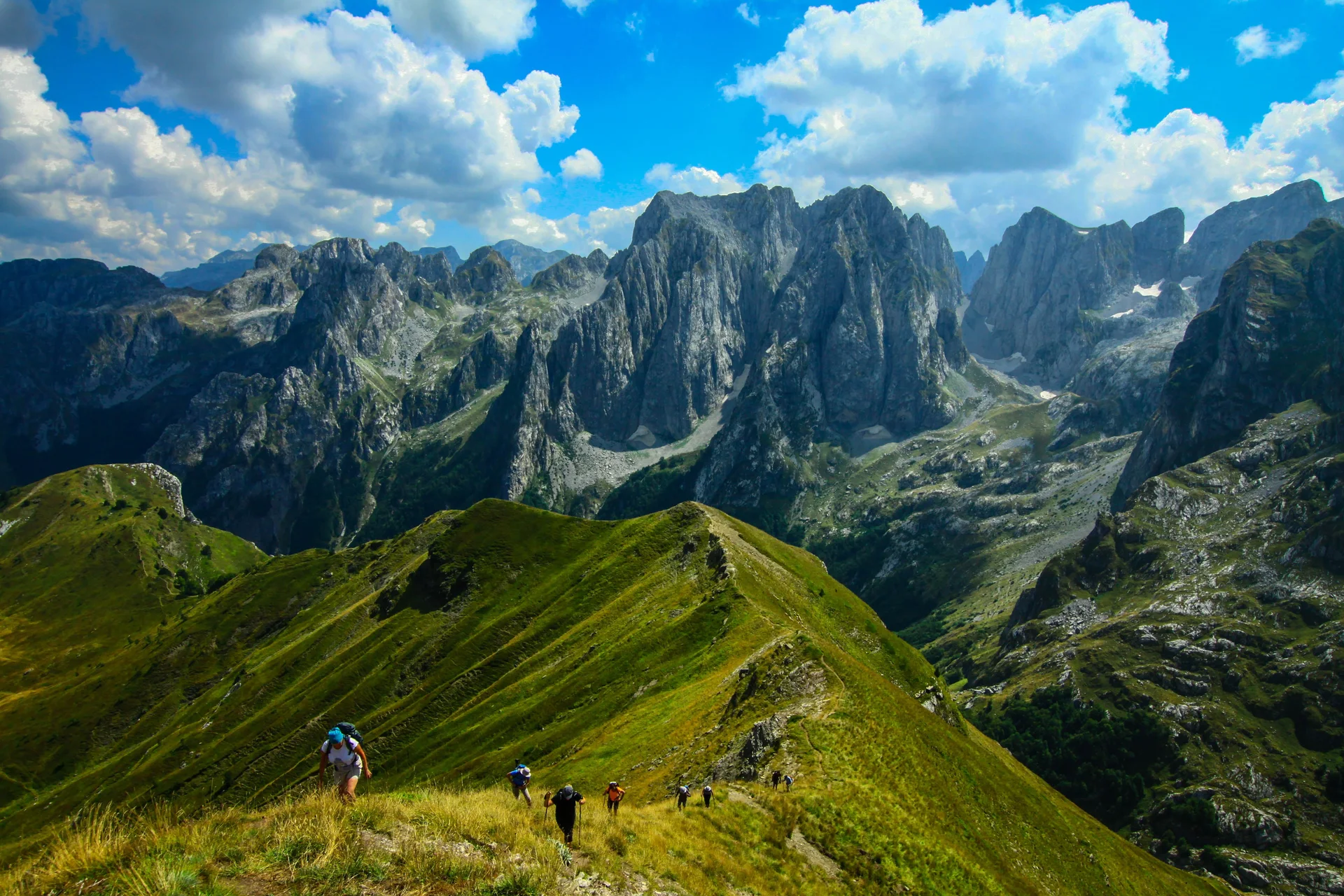 Hikers climbing a grassy mountain slope beneath the limestone peaks of the Accursed Mountains.